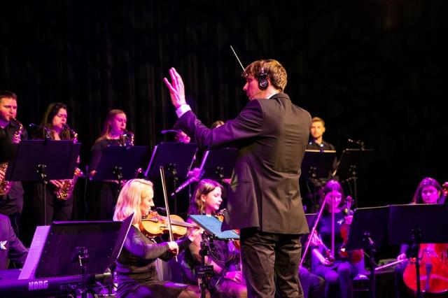 Zdjęcie przedstawia dyrygenta oraz orkiestrę grającą koncert